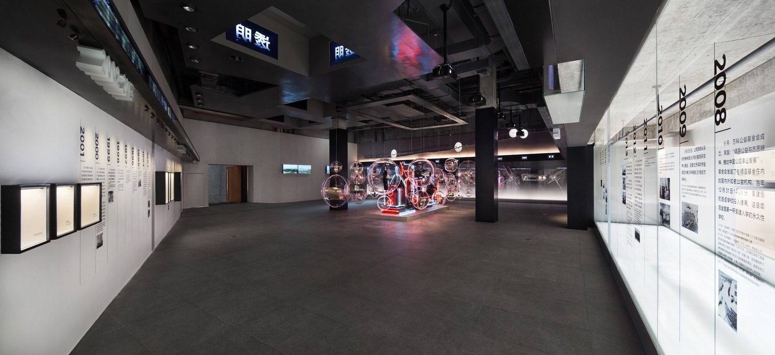 历史馆是一个长卷式的展示, 从万科的前身开始, 到一直演变和发展的重要大事记。 我们以阶段性的年份来划分空间, 平面的文字和立体的展示柜交互配合, 形成一个有趣的阅读体验, 在大事记的展墙上方, 我们设计了一个环状的LED屏, 循环播放代表万科理念的文字。空间中间部分摆放了艺术家周文斗的作品, 他利用许多大小不一的透明塑料球把代表万科历史的物件重新包装起来, 形成一个富有历史感的艺术作品。